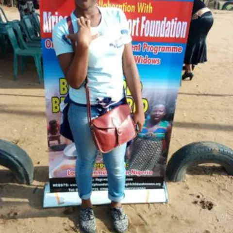 A woman in a green checked apron and hairnet making dough rings (donuts or puff-puff) on a white table outdoors.