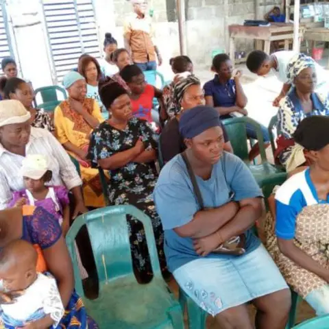 A full view of women of all ages packed into green plastic chairs under a striped canopy tent at an outdoor community programme.