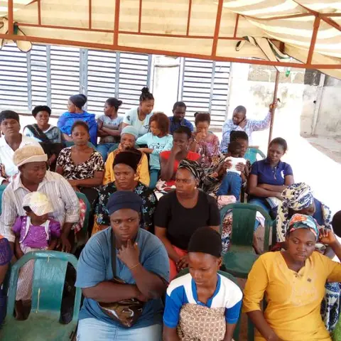 A wide-angle view of women seated in rows under a striped canopy tent, with colourful clothing visible among the crowd.