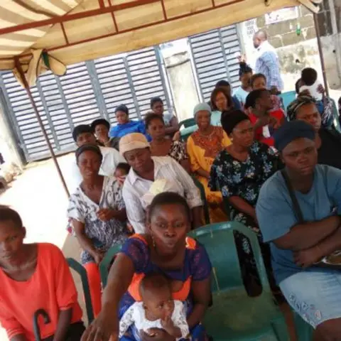 Women in rows, several holding babies and toddlers, seated at an outdoor community empowerment event under a canopy.