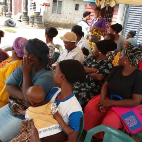 Women seated at an outdoor event under a canopy, with parked vehicles visible in the background along a dirt road.