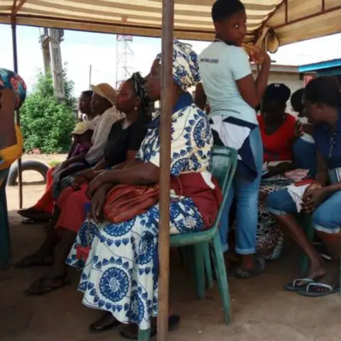 Women seated under a canopy tent, with a woman in a green patterned headscarf listening in the foreground.
