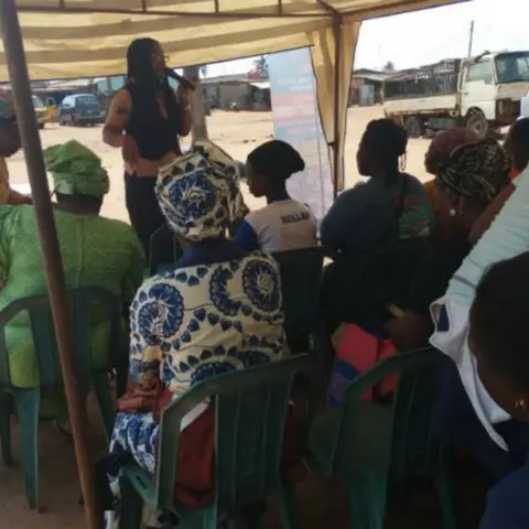 A woman in dark clothing stands addressing a large seated audience of women under a canopy tent at a roadside event.