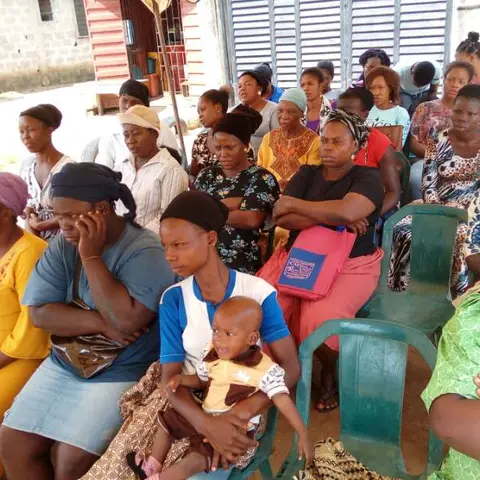 A large outdoor audience of women and children; a mother in the foreground holds a baby in blue and orange patterned fabric.