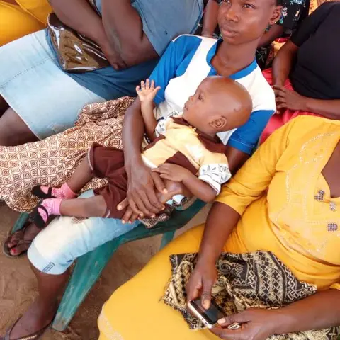 A close-up of a young woman holding a sleeping baby on her lap, seated next to a woman in a yellow dress.