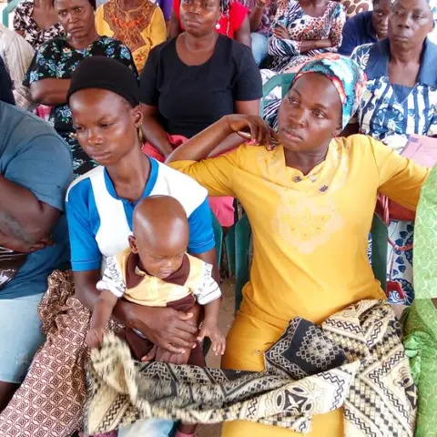 A side view of women seated in rows, some holding babies and bags, at an outdoor empowerment programme.