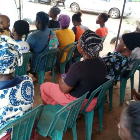 Women seated in rows of green chairs, with young men in jeans in the foreground, at an outdoor community event.