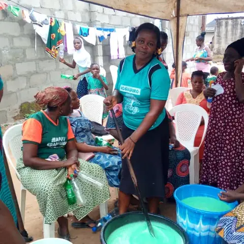 A woman in a teal shirt stirs large buckets of green liquid soap at an outdoor soap-making demonstration, with the AFSF banner visible above.