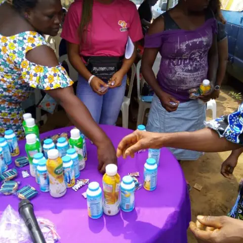 Community members reach towards a table of nutritional drinks and health products at an outdoor distribution event.
