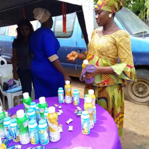 A woman in a yellow and gold dress receives a nutritional drink from a table of health products at an outdoor outreach event.