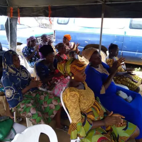 Women under a canopy tent applaud with raised hands in celebration at an outdoor community empowerment gathering.
