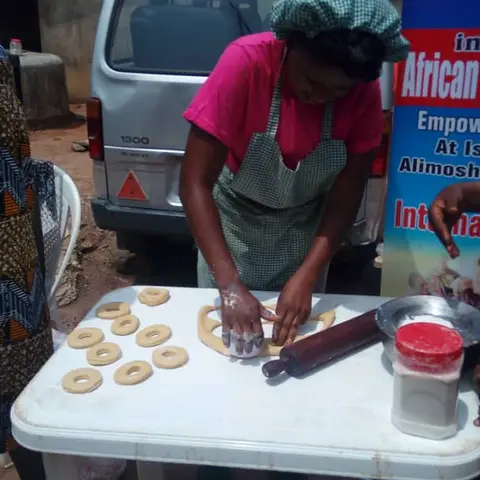 A woman in a pink top, checked apron, and headscarf shapes dough rings on a white table at an outdoor baking skills training, with an AFSF banner partially visible.