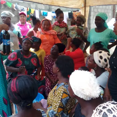 A woman in a teal Igidaduro t-shirt demonstrates soap-making by stirring green liquid in a large bucket, surrounded by women watching under a canopy.