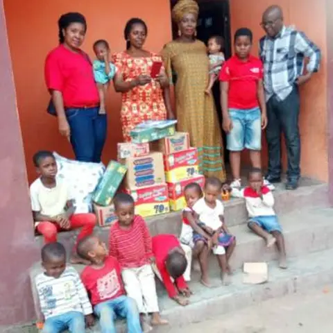 Adults and children standing on the steps of an orange building with stacked donated supplies during a mission outreach visit.