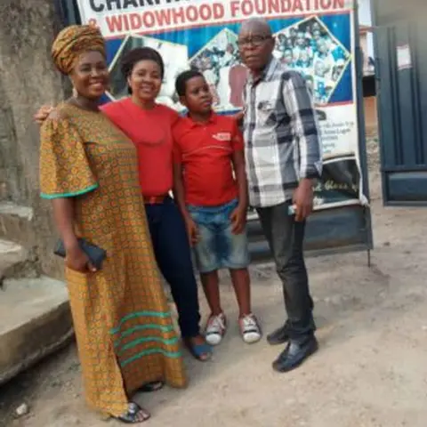 Four individuals – two women, a boy, and a man – posing in front of the Charitas Orphanage and Widowhood Foundation banner.