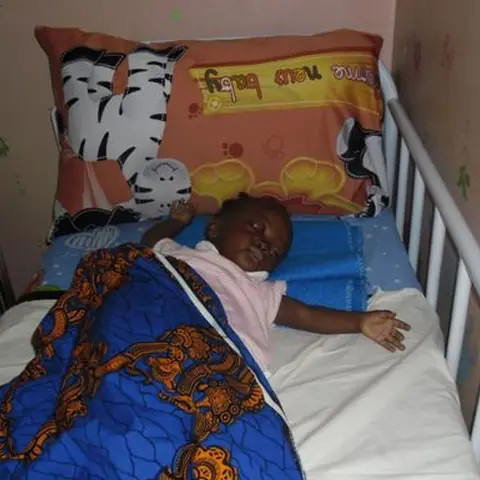 A young child lying in a cot covered with a blue patterned fabric in a small room with floral wall stickers.