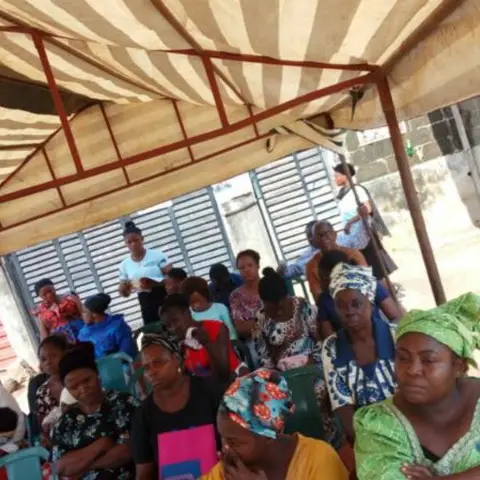 Women and children seated under a striped canopy tent at an outdoor community meeting in Nigeria, viewed from the side.