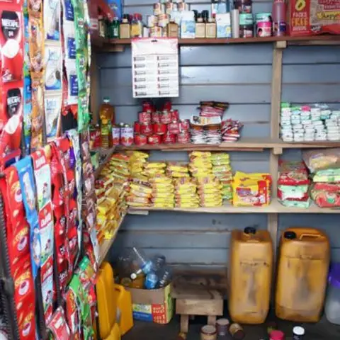 The shelves of a small community shop stocked with tinned goods, snack sachets, cooking ingredients, and yellow jerricans.