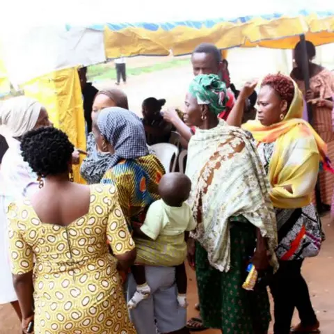 Women and community members gathering and milling around under a yellow tent at an outdoor distribution event.
