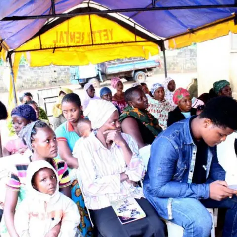 Women and young people seated under a yellow rental canopy tent, some looking at their phones, at an Africa Mission outreach event.