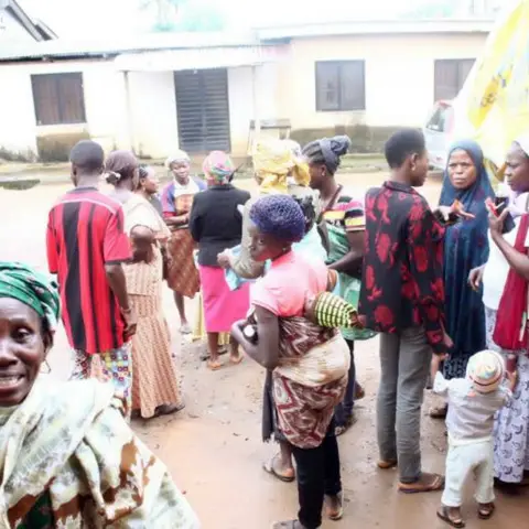 Community members queuing near a yellow tent at an outdoor distribution event; an elderly woman smiles warmly in the foreground.