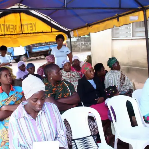Women seated in white plastic chairs under a yellow canopy tent, listening to a speaker at an Africa Mission community programme.
