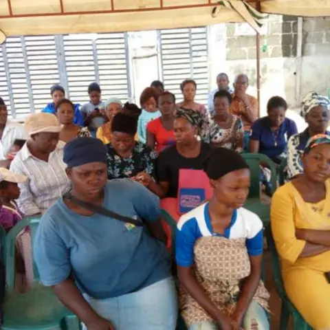 A packed crowd of women and children in green plastic chairs under a canopy, listening attentively at an outdoor empowerment programme.