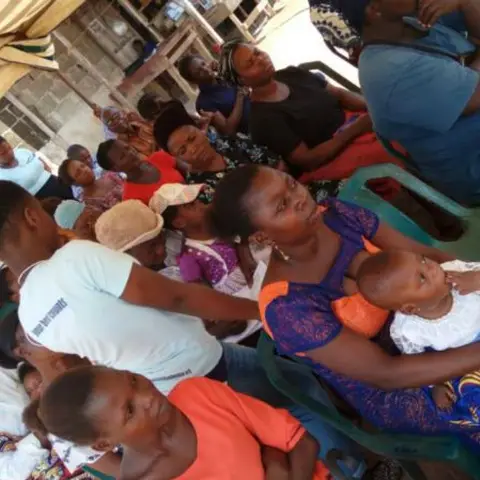 Women and children crowded under a canopy tent; a woman in a blue patterned dress holds a baby in the foreground.