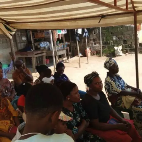 Women and children seated under a striped canopy tent, with a community organiser standing at the back left.