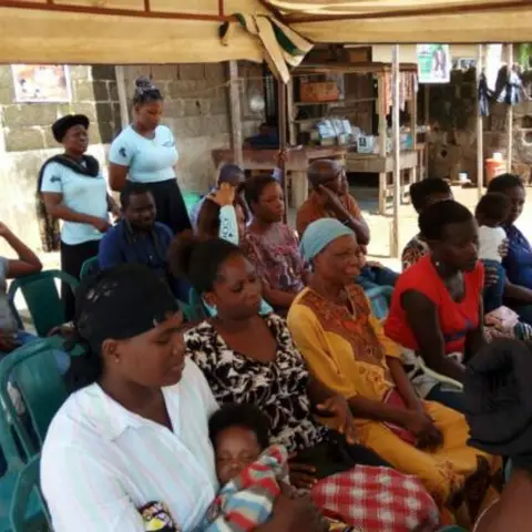 A woman in a white shirt stands at the back while women and children are seated in green plastic chairs under a canopy tent.