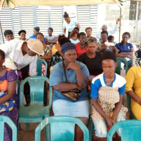 A large crowd of women seated in green plastic chairs under a striped canopy tent, some holding babies, at an outdoor empowerment event.