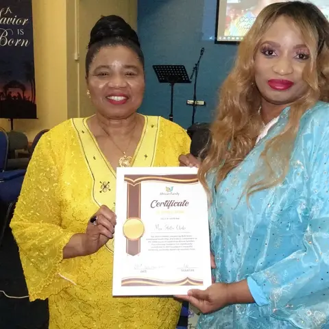 A man in a blue suit and cap presenting a certificate to a woman in a light blue dress, while another woman in yellow looks on.