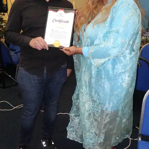 A close-up of the presenter in a light blue dress speaking to the audience, holding a certificate folder.