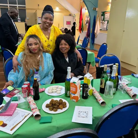 Three women posing together, one in a yellow dress, one in light blue, and one in a black coat.