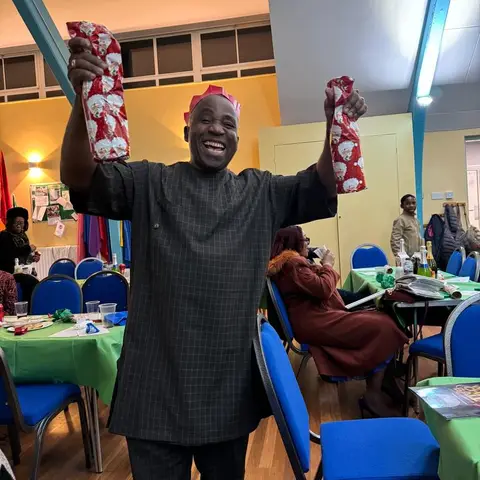 Three women standing together in the hall, smiling, with tables and blue chairs in the background.