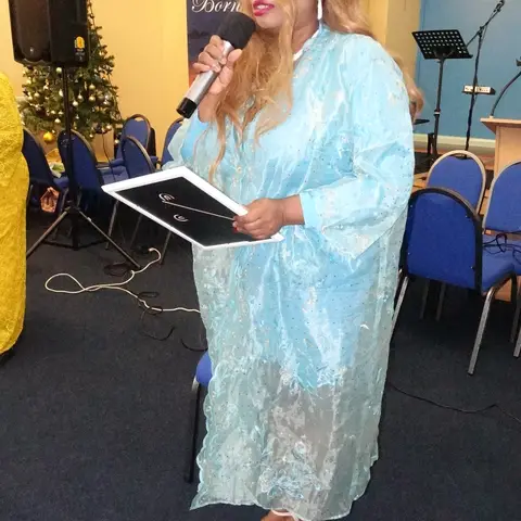 A woman in a light blue dress speaking into a microphone while holding a certificate folder, next to a Christmas tree.