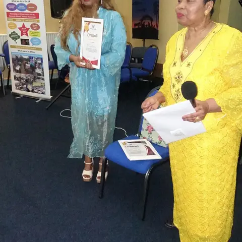 A woman in a bright yellow dress speaking with a microphone while standing next to a woman holding a certificate.