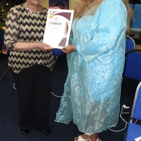 A woman in a black and gold zigzag patterned top receiving a certificate of appreciation from the presenter.