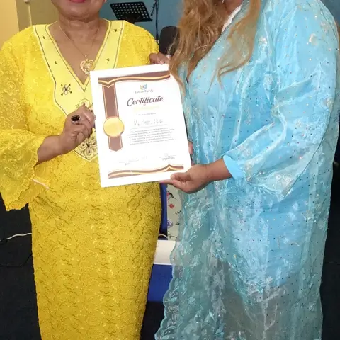 A woman in a yellow dress proudly displaying her certificate of appreciation alongside the event host.