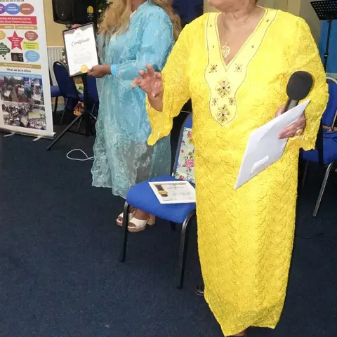 A man in a black shirt smiling as he receives a certificate from the presenter in a light blue dress.