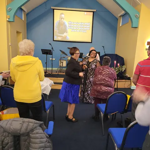 Women dancing joyfully in a church hall during a gathering, with lyrics projected on a screen in the background.