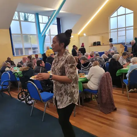 A woman walking back to her seat with a plate of food, with the busy community hall in the background.