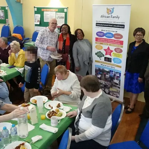 Community members and volunteers posing for a group photo next to an African Family Support Foundation banner during a meal.