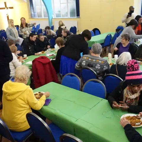 A wide view of a community hall with guests seated at long tables with green tablecloths, enjoying a meal and conversation.