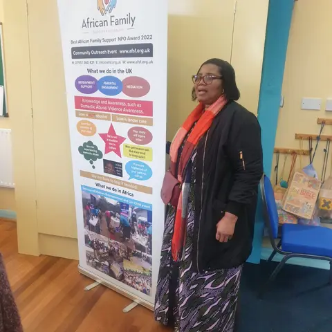 A woman standing next to the African Family Support Foundation banner, which details the organization's services and awards.