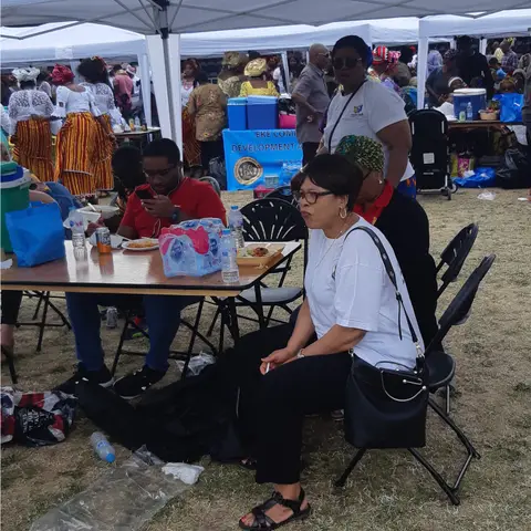 A candid shot of attendees relaxing under a white gazebo at an outdoor festival, with a woman in a white t-shirt and black pants sitting in the foreground.