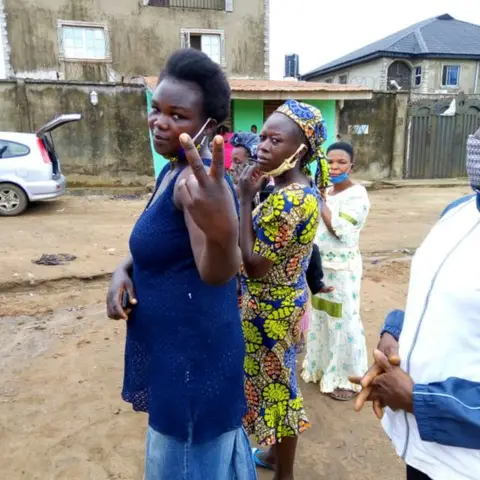 Three community women wearing face masks outdoors during a COVID-era food distribution; one woman gestures a peace sign.