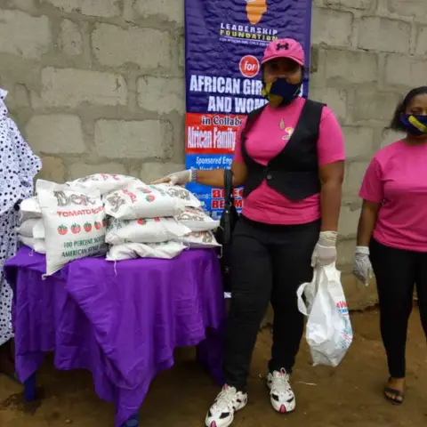 Two foundation volunteers in pink shirts and face masks standing with bags of food beside the Leadership Foundation for African Girl Child and Women banner.