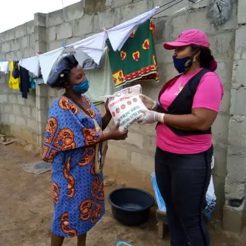 A foundation volunteer in pink hands a bag of Best Tomato Rice to a woman in a blue patterned dress during a home-visit food distribution.