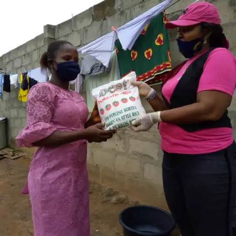 A foundation volunteer in pink hands a bag of rice to a woman in a pink lace dress at an outdoor food distribution event.